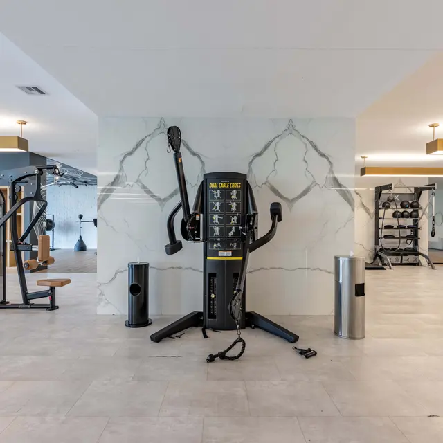 A spacious and modern gym interior featuring various exercise machines, including a multi-station unit. The floor is tiled, and the walls have light colors with decorative elements. The lighting is bright, with fixtures suspended from the ceiling.