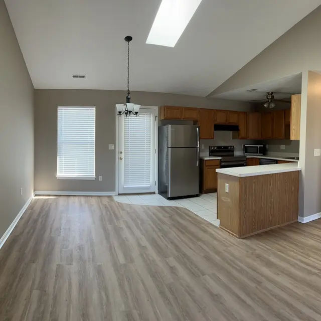 A spacious living area with a kitchen in the background, featuring wooden cabinets, stainless steel appliances, and large windows allowing natural light to enter.