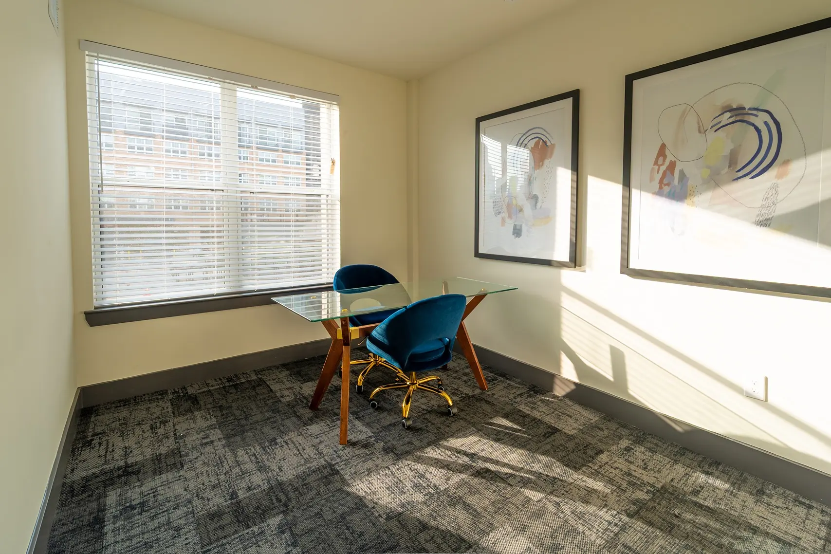 A modern office space with a glass table and two blue chairs. The room features large windows with blinds and two framed abstract art pieces on the walls.