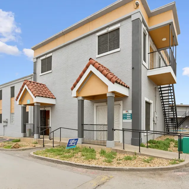 Modern two-story apartment building with decorative roof accents and a staircase, surrounded by greenery and parking area.