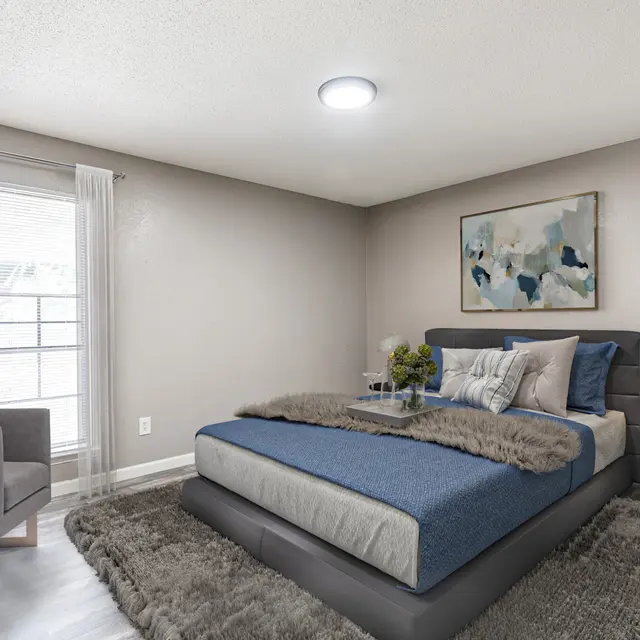 A modern bedroom featuring a comfortable bed with a blue cover, surrounded by a cozy rug and an accent chair. Large window allowing natural light, with soft curtains and minimal decor.