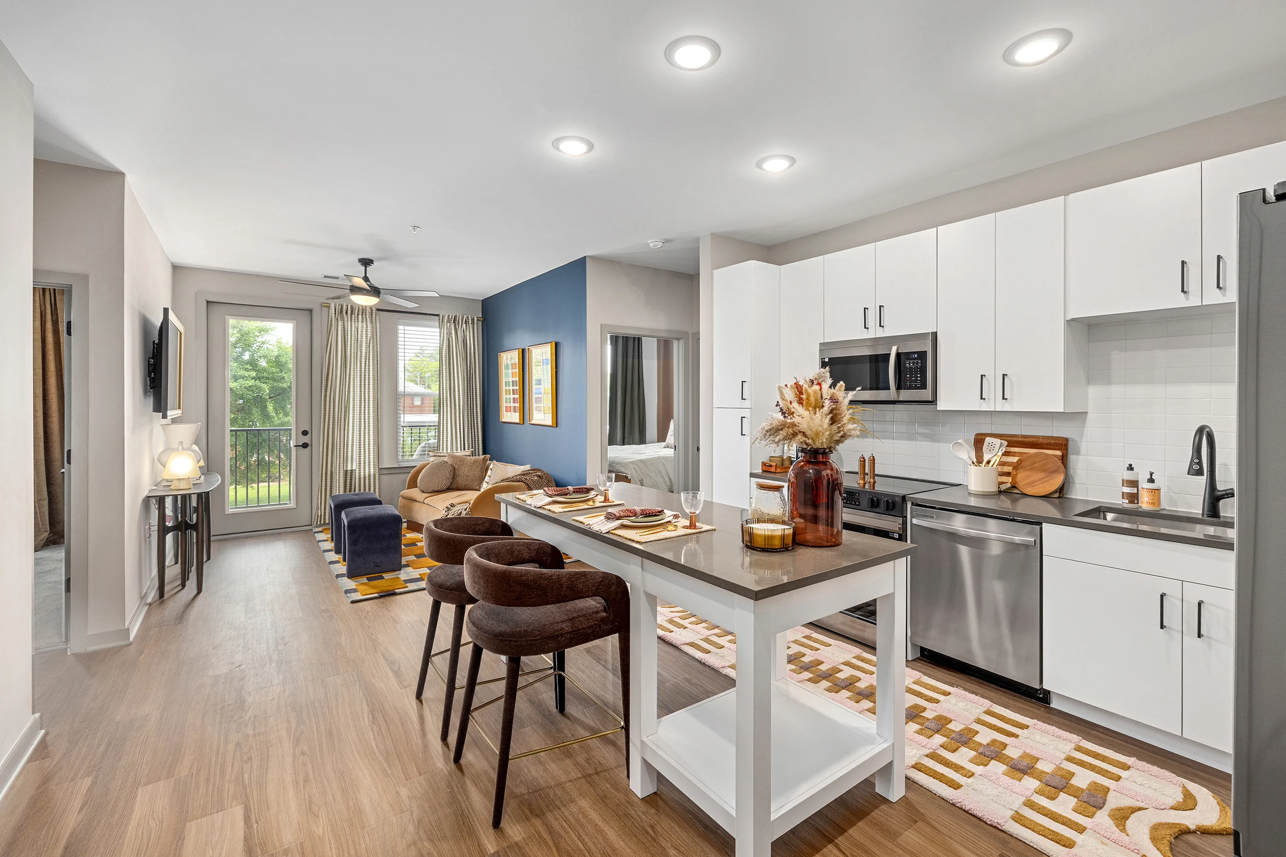 Modern Kitchen and Living Room A modern open-concept kitchen and living area featuring white cabinets, stainless steel appliances, a comfortable brown armchair, and a dining table with four stools. Large windows open to a view of greenery outside.