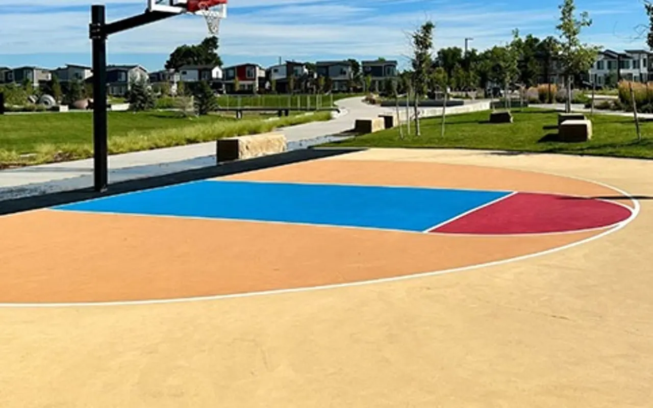 A vibrant outdoor basketball court featuring a blue and red paint scheme on a yellow surface, with a basketball hoop in the foreground and green grass surrounding the area.