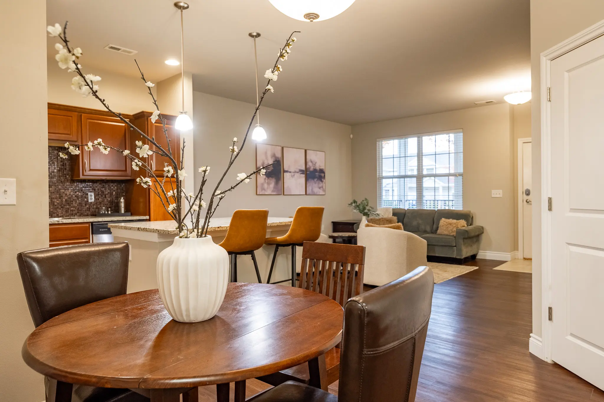 A modern apartment interior featuring a dining area with a round table and a vase of branches, kitchen in the background, and a cozy living room visible in the distance.