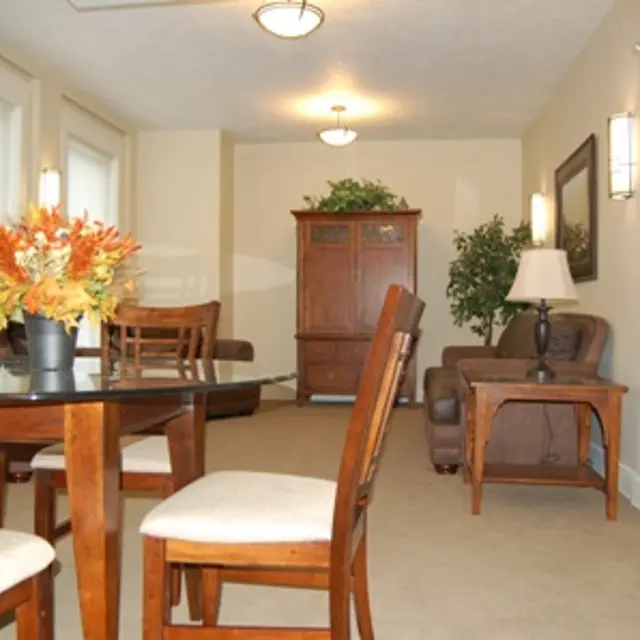 Cozy Living and Dining Area A cozy living room and dining area with a glass table, wooden chairs, and a brown couch. The space is bright with windows and decorative plants.