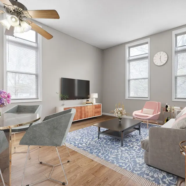 A modern living room featuring a light grey wall, wooden floor, a cream-colored sofa, and a glass dining table surrounded by silver chairs. A TV is mounted on the wall with a wooden TV stand below. There is a light pink chair near the windows, and a rug with a blue design on the floor. Fresh flowers in a vase are placed on the dining table, and a clock is hanging on the wall.