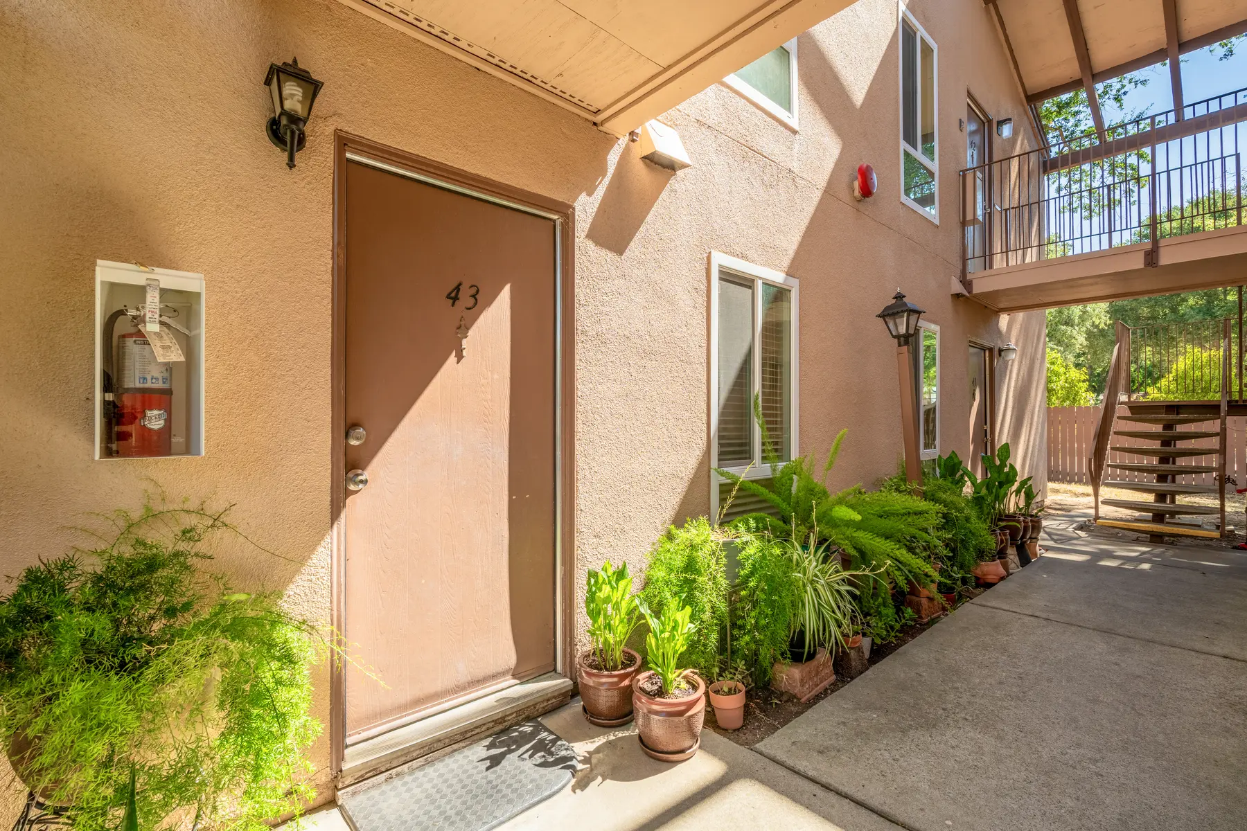 An apartment entrance with a brown door numbered 43, surrounded by potted plants. The setting features a wall-mounted fire extinguisher, and a staircase leading to the upper floor in the background.