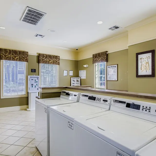 A bright and clean laundry room featuring multiple washing machines and a counter space. The walls are painted in a light olive shade, with large windows that have decorative valances and blinds. There are several notices and framed artwork on the walls, and the room is well-lit with ceiling lights.