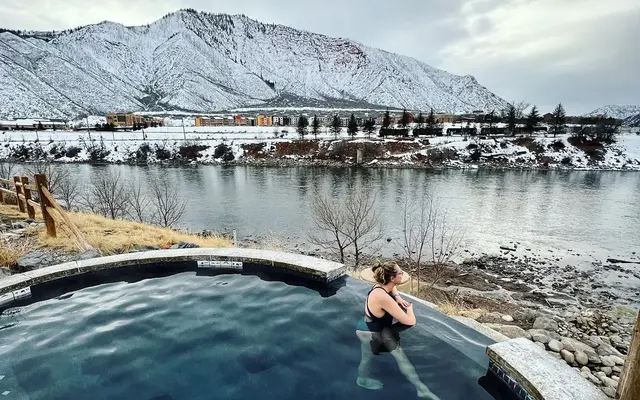 A woman enjoying a hot spring surrounded by snowy mountains and a river.