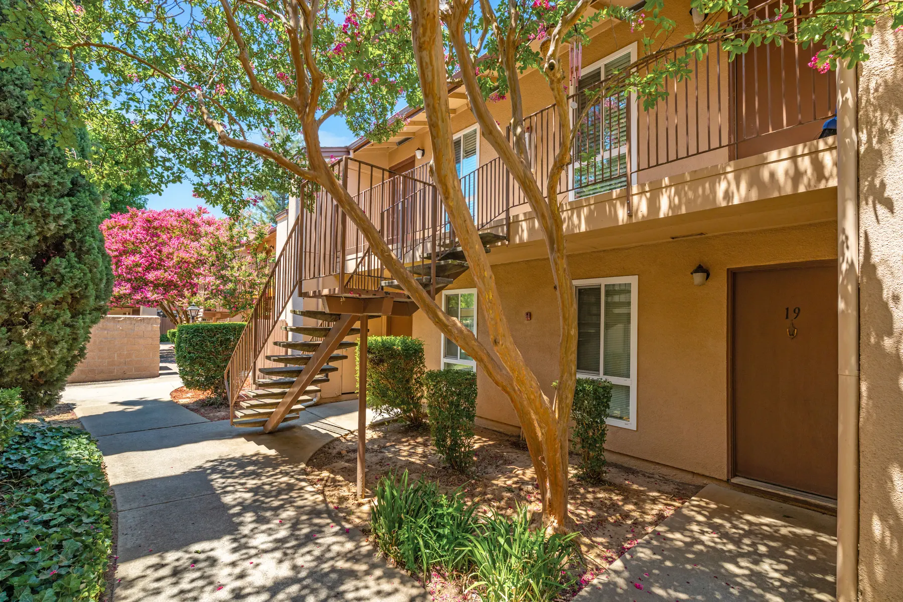 A pathway leading to an apartment complex with stairs and lush greenery, featuring a large tree and colorful flowers in the background.