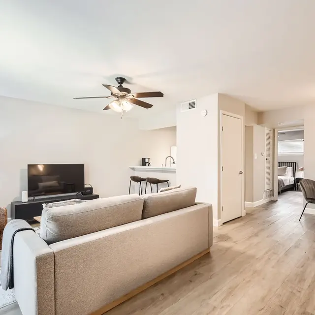 A modern apartment interior featuring a living area with a sofa, TV, and a dining space with chairs. Bright lighting and wooden flooring enhance the spacious feel.