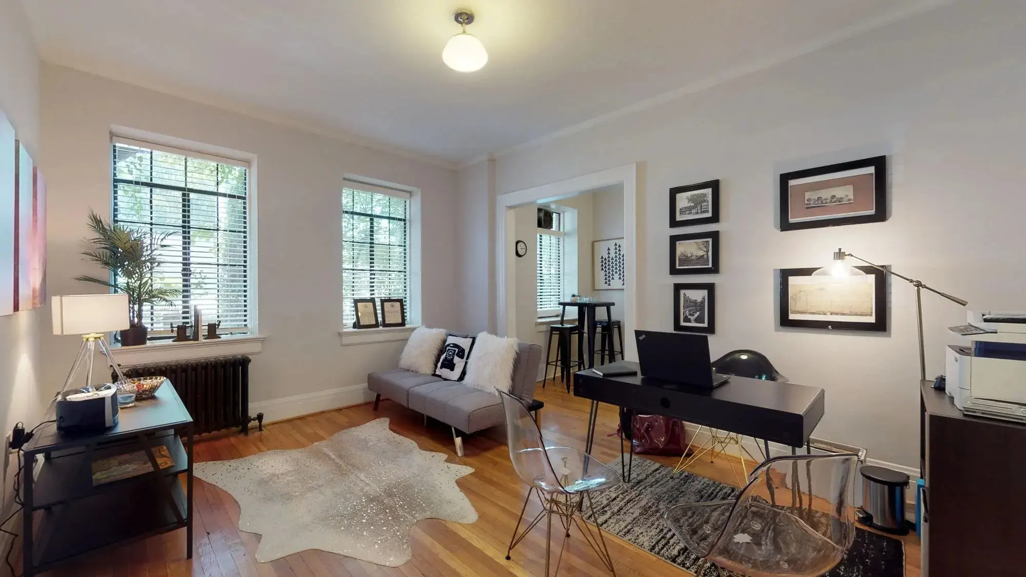 Cozy Apartment Living Room A cozy living room with a sofa, a desk, and several windows letting in natural light. Decorative items adorn the walls, and a soft area rug is on the floor.