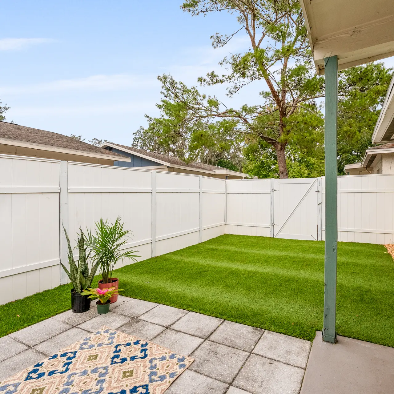 A backyard patio featuring artificial grass and white wooden fencing, with a small decorative tile area and potted plants.