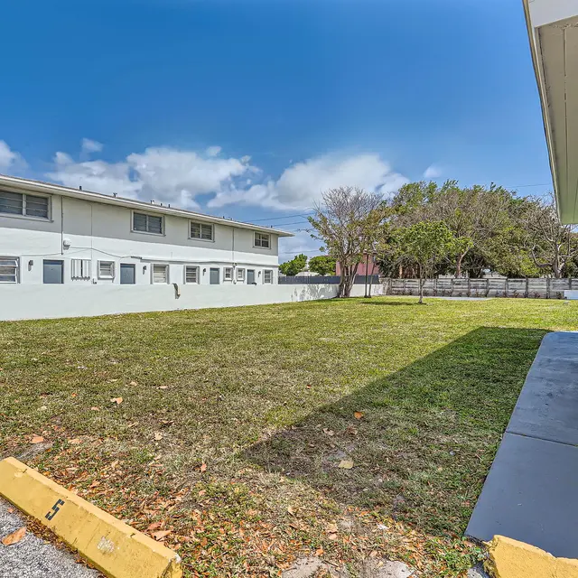 A view of an apartment building with an open lawn area in front, surrounded by trees and blue sky.