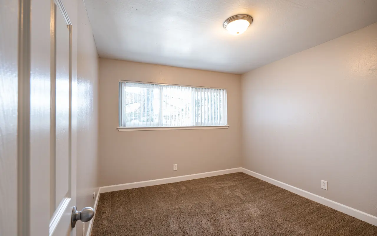 A vacant room with beige walls and carpet, featuring a single window with blinds, and a light fixture on the ceiling.