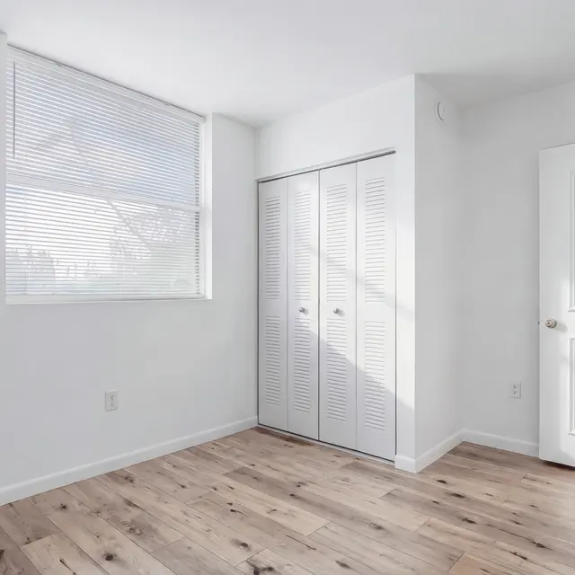 Empty room with white walls and a wooden floor, featuring a closet with sliding doors and a window with blinds