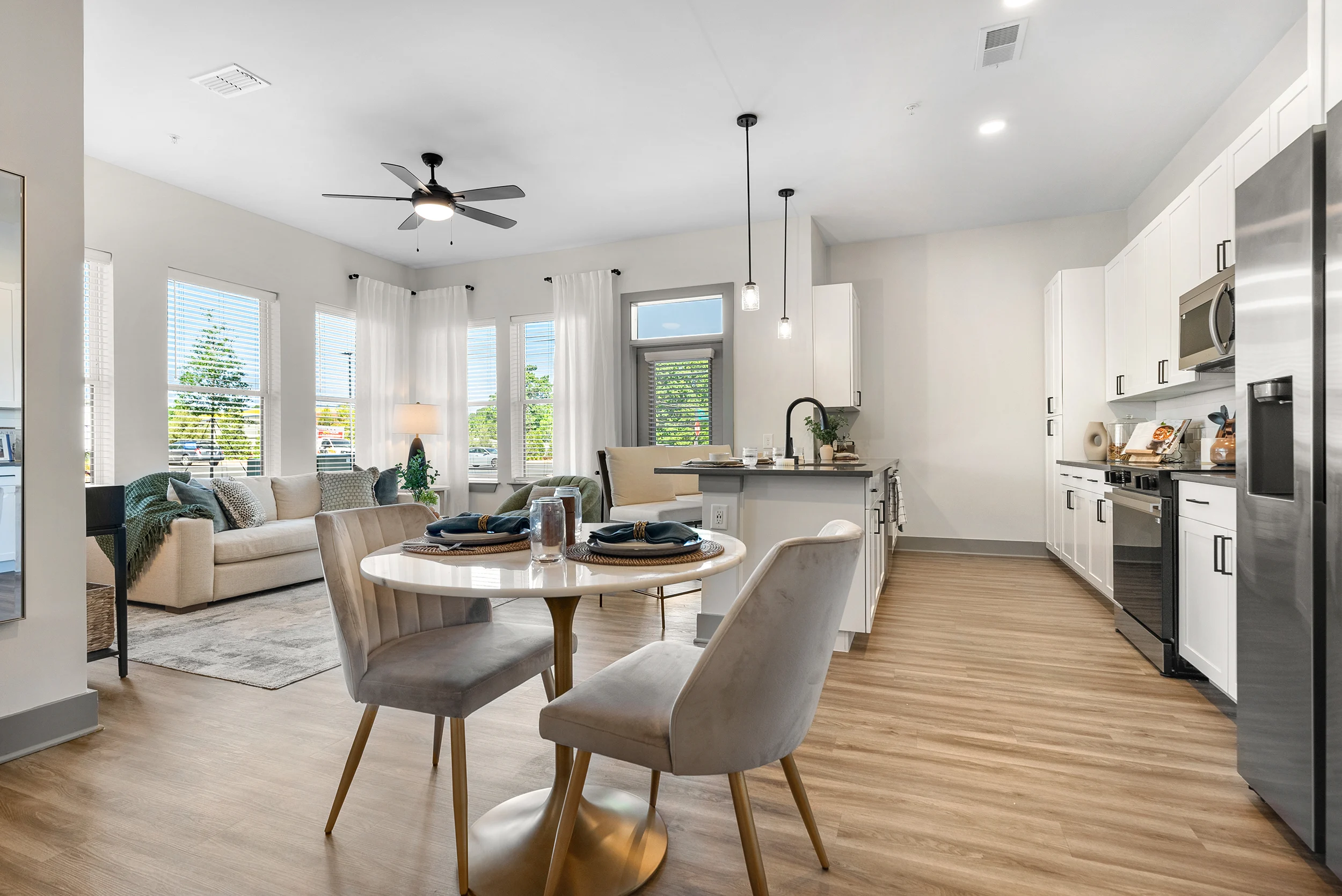 A modern apartment interior featuring a kitchen and living room area. The kitchen incorporates white cabinets, stainless steel appliances, and a circular dining table with two chairs. Natural light is streaming through large windows.