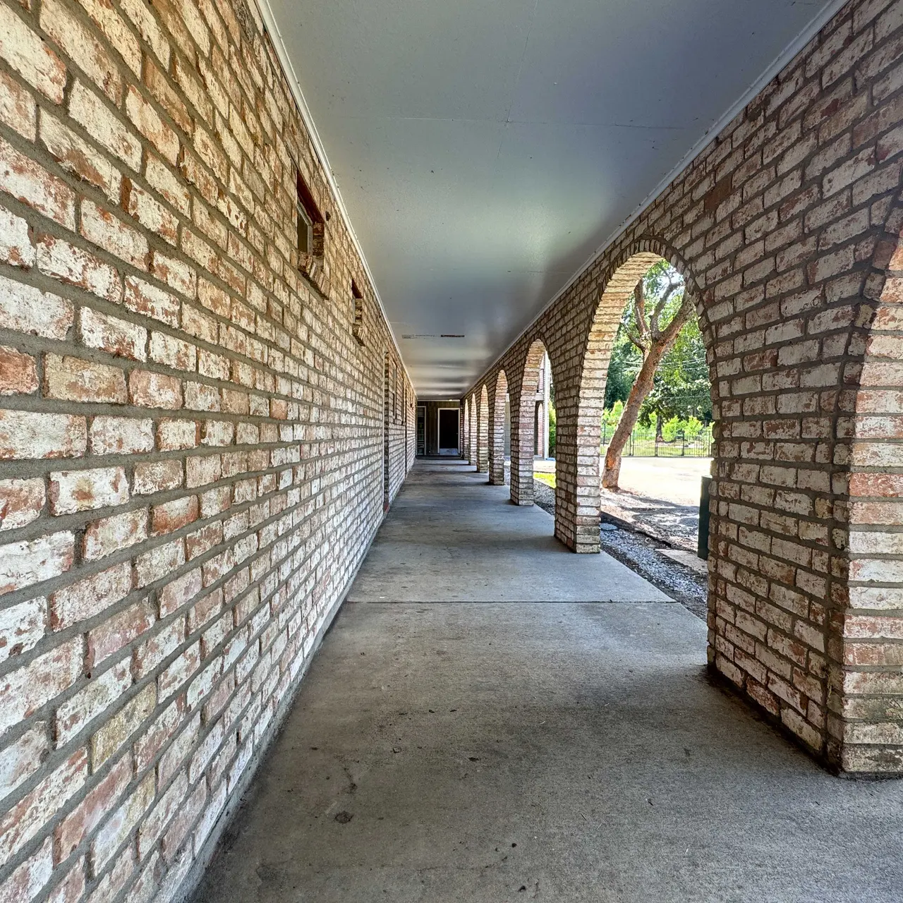 A long, narrow hallway with brick walls and arched openings leading to a green area outside.