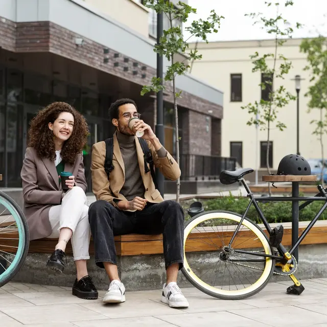 A young couple sitting on a bench in a modern outdoor setting with two bicycles beside them. The woman has curly hair and is smiling while holding a drink. The man is wearing glasses and drinking from a cup, looking relaxed. They are dressed stylishly in casual outfits, and there are green trees in the background.