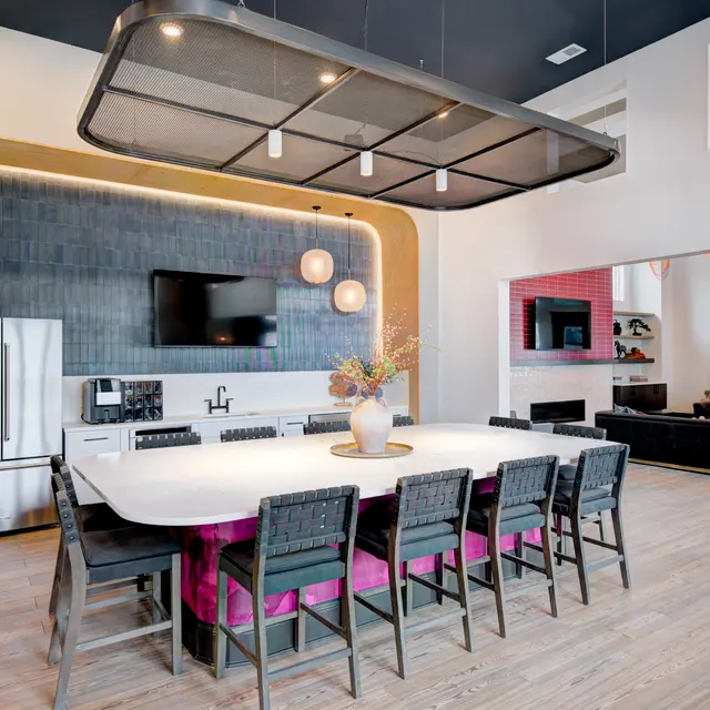 A modern kitchen with a large dining table, stylish chairs, and appliances including a refrigerator and coffee machine. The decor features contemporary lighting and a sleek design with wood flooring.