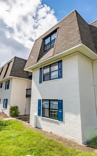 A two-story apartment building with a white exterior and blue shutters, set against a backdrop of partly cloudy skies. The building has a grassy front yard with trees and landscaping.