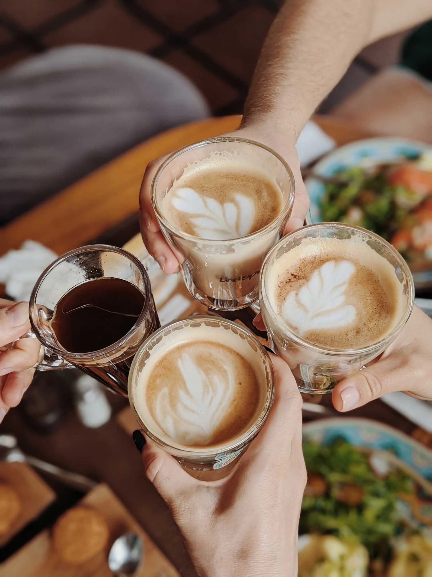 Coffee Toast Four hands holding glasses of coffee with latte art, a wooden table with food in the background.