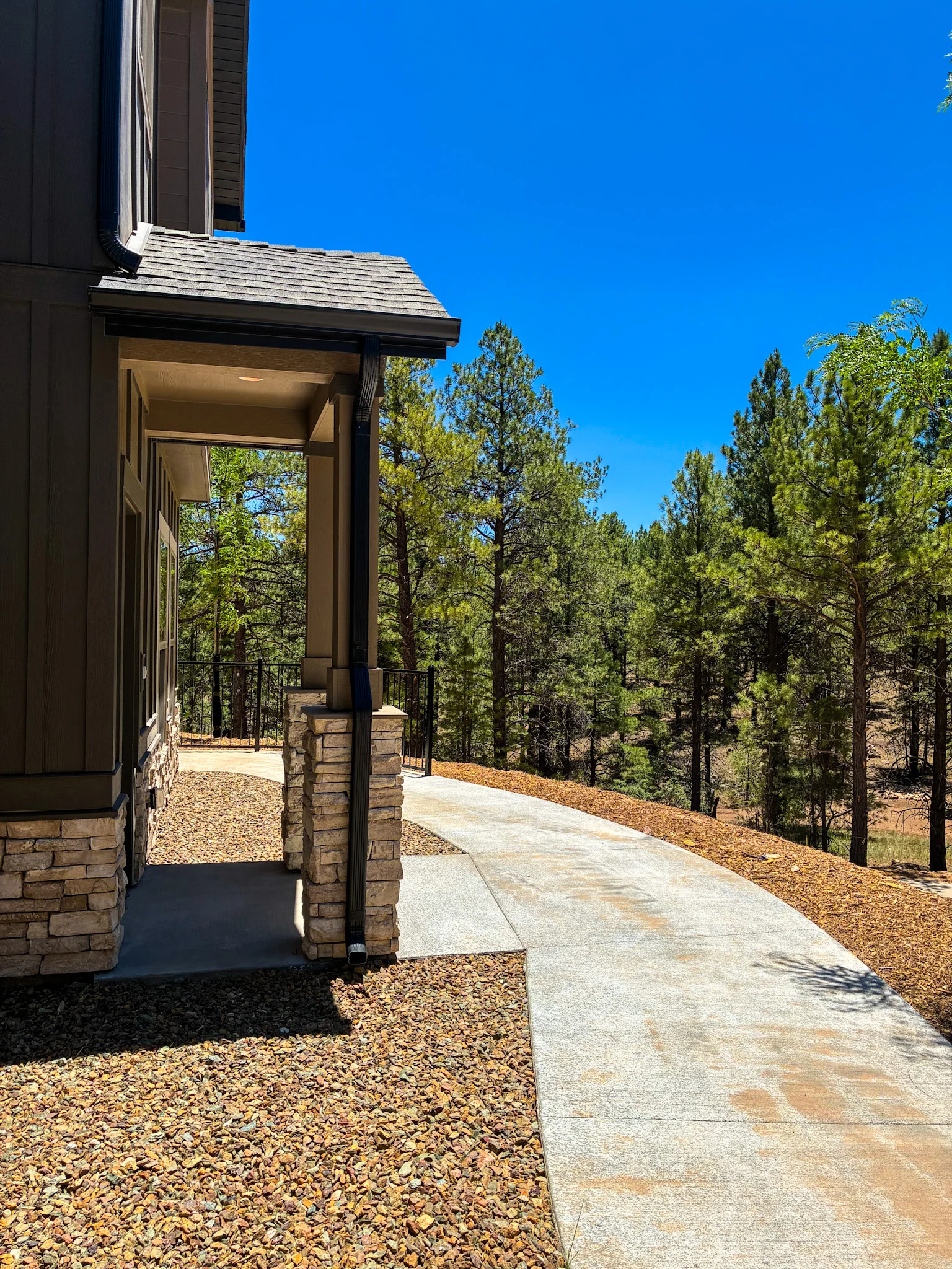 A winding pathway leading alongside a house, surrounded by tall trees under a clear blue sky.