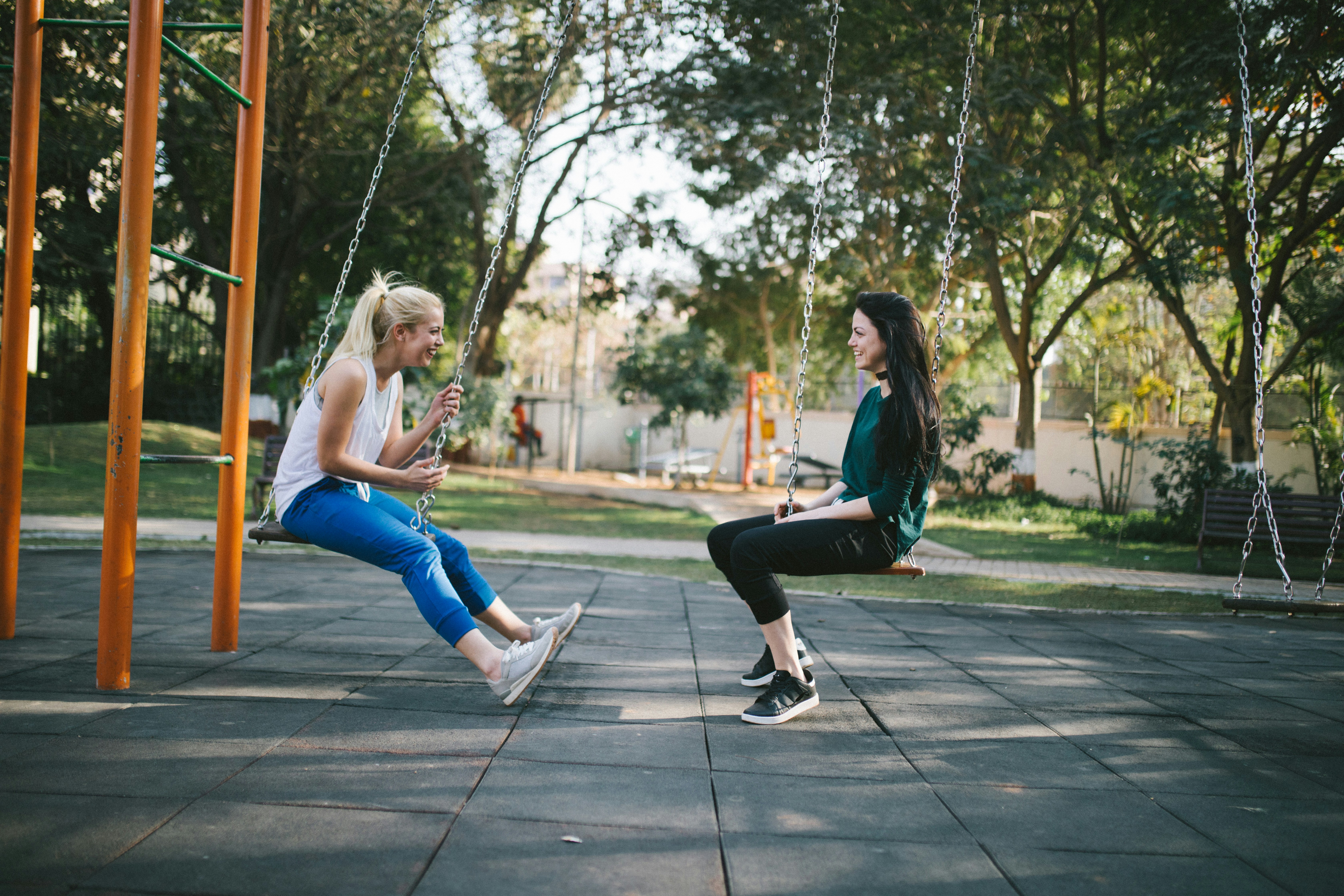 Two women sitting on swings, enjoying a sunny day in a park with trees in the background.