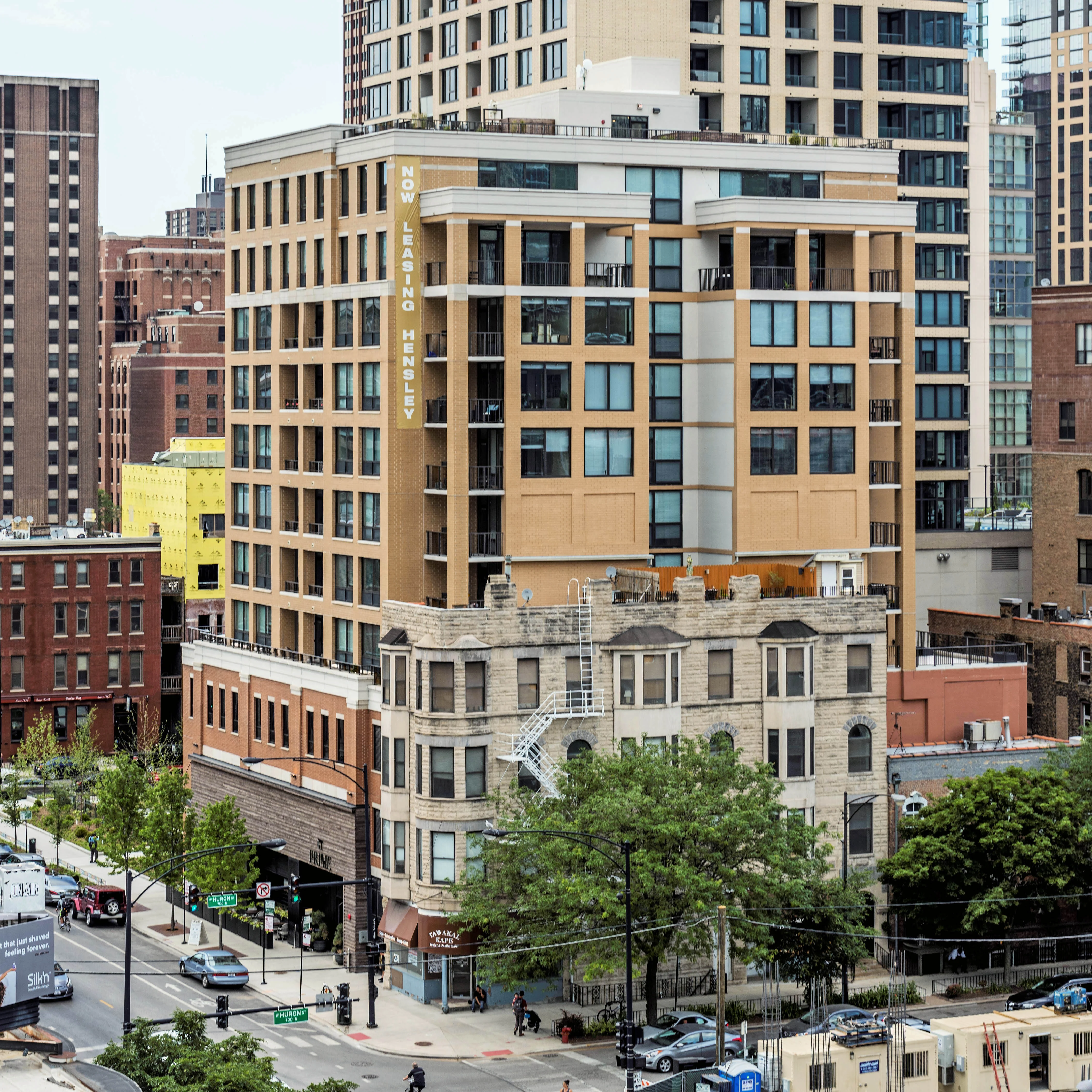 Urban Landscape with Historic and Modern Buildings A view of a city block featuring a mix of modern and historic architecture. The foreground shows a tree-lined street with buildings of various heights and designs, including a newer high-rise and an older brick structure. There are cars parked along the street and people walking.