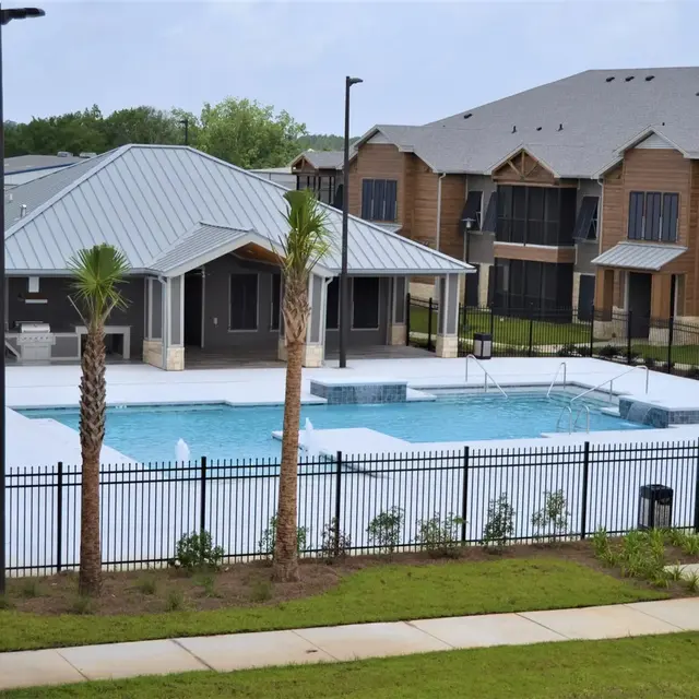 A spacious pool area surrounded by green grass and palm trees, featuring a modern clubhouse, lounge chairs, and a fenced perimeter.