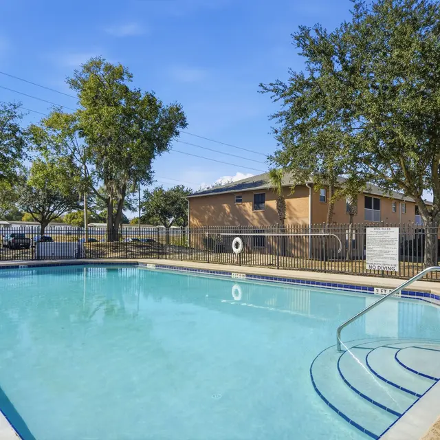 Clear swimming pool surrounded by trees and a fence at an apartment complex, with a few residential buildings in the background.