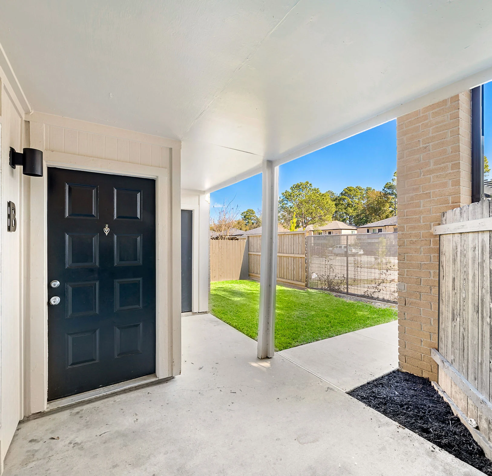 A view of an entrance with a black door on the left side, a concrete walkway, and a fenced yard visible in the background.
