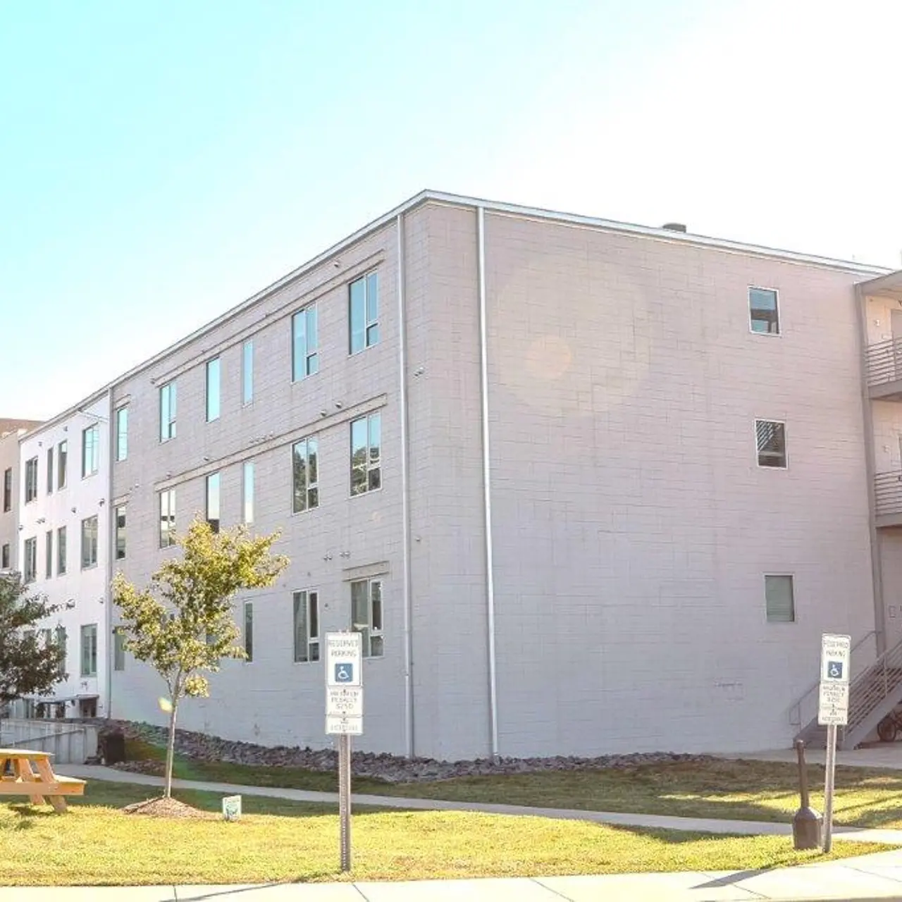 A modern building exterior with large windows, a grassy area in front, and an adjacent fire escape. Two small trees are visible near the sidewalk.