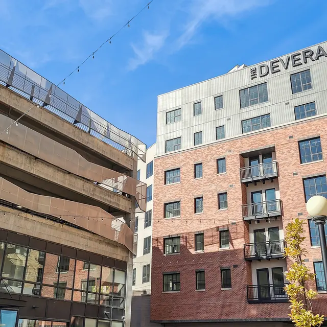 Urban Apartment Building - The Deveraux A street view of a modern, brick apartment building named The Deveraux, flanked by a concrete structure on the left. The sky is clear with a few clouds.