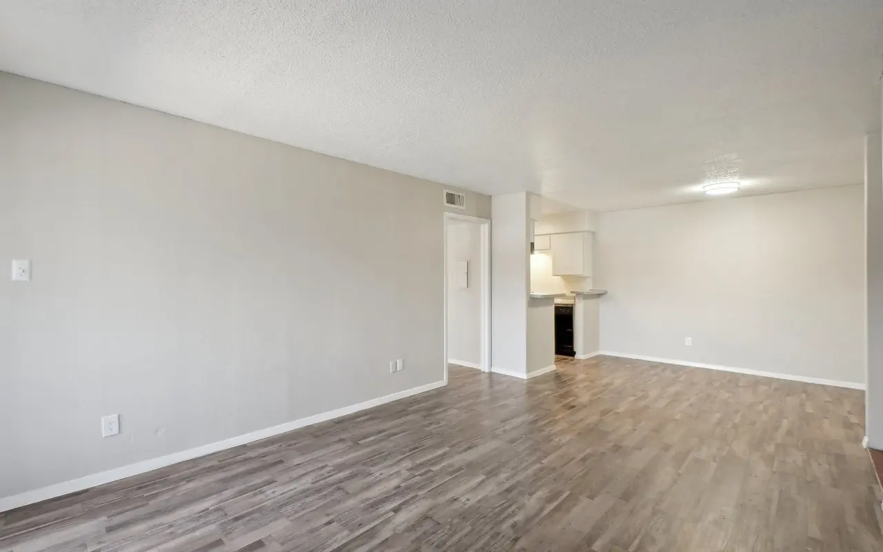 A spacious empty living room with light-colored walls and wood flooring. The kitchen area is visible in the background.