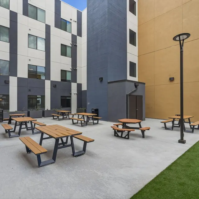 An outdoor seating area featuring multiple wooden picnic tables on a concrete surface, surrounded by modern buildings in light and dark colors, with a patch of green grass and a lamp post.
