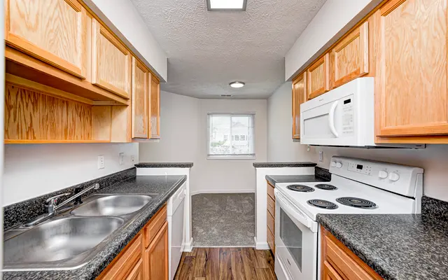 A modern kitchen featuring wooden cabinets, a double sink, and white appliances including a stove and microwave, with a window providing natural light.
