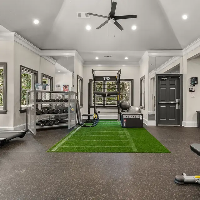 A modern gym interior featuring various fitness equipment including treadmills, dumbbells, and a multi-station exercise machine, with an artificial turf area for training.