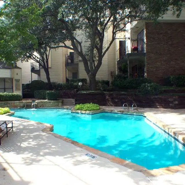 A serene apartment complex pool area featuring a blue pool, surrounded by lush greenery and lounge chairs. The backdrop includes apartment buildings with balconies and shading trees.
