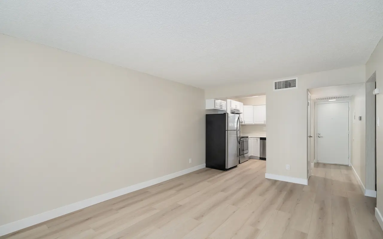An empty living room with light wood flooring, featuring a black refrigerator and kitchen area in the background. Walls are painted a neutral color, creating a spacious and airy feel.