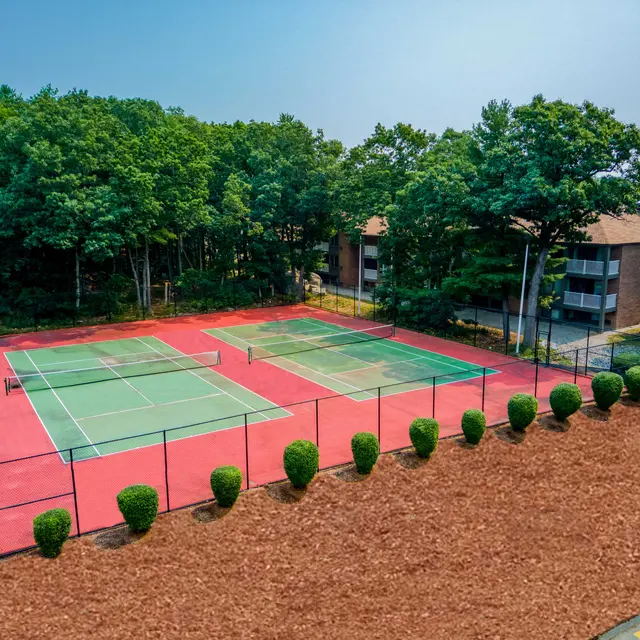 Two outdoor tennis courts surrounded by greenery and apartment buildings, featuring red and green court surfaces.