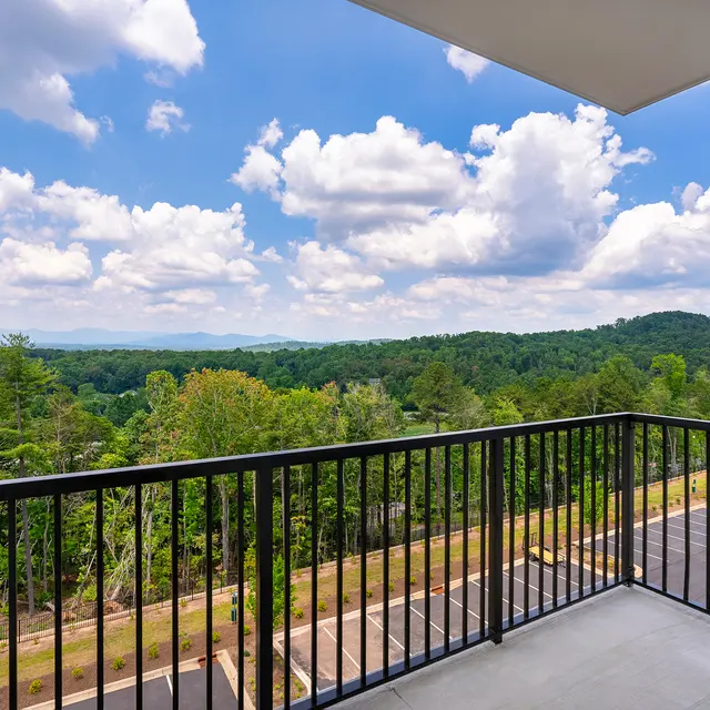 A balcony with a black railing overlooking a lush green landscape and blue sky with fluffy white clouds.