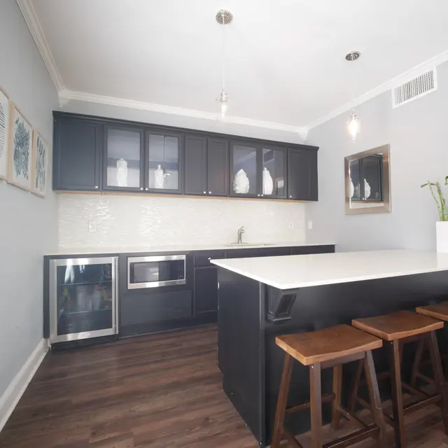 A modern kitchen with dark cabinets and a white countertop. A small dining area with wooden stools is positioned at the kitchen island. Light fixtures hang from the ceiling.