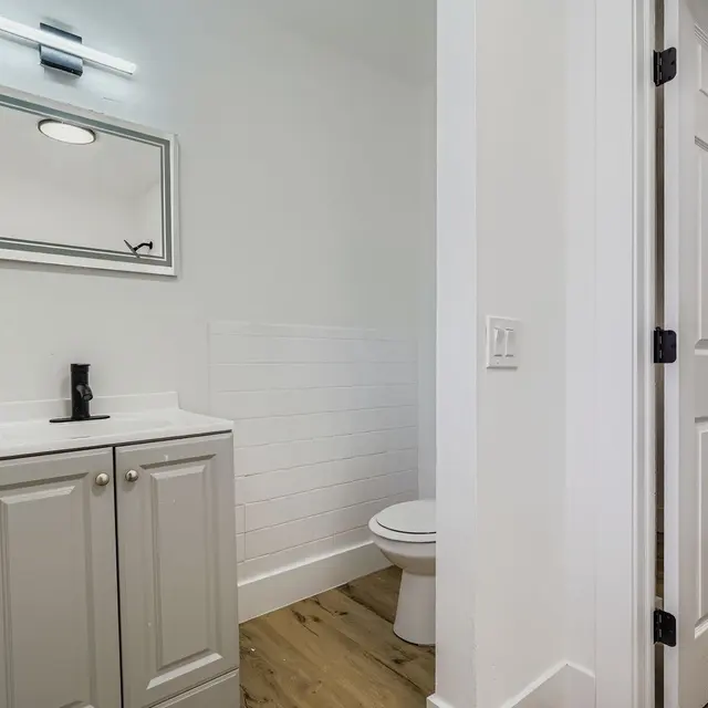 A modern bathroom featuring a white and gray color scheme, including a sleek vanity with a mirror and a toilet. The flooring is wooden, and the walls are painted white.