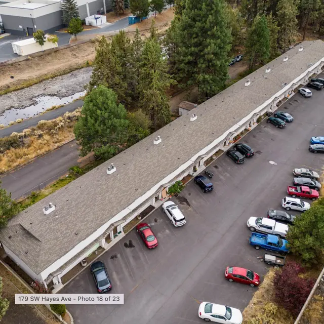 Aerial view of a residential building with cars parked in front and trees surrounding the area. A stream is visible beside the property.