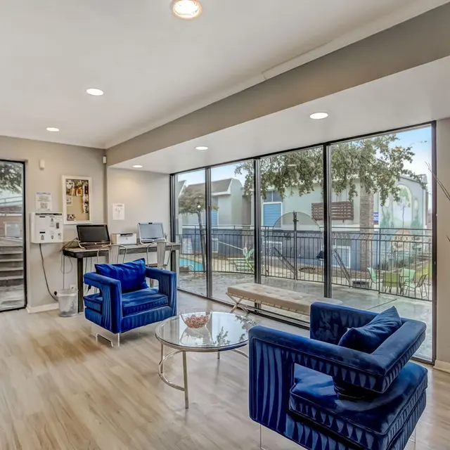 A modern lobby interior featuring blue upholstered chairs and a glass table with a decorative bowl. Large windows provide natural light and a view of an outdoor area with trees and steps.
