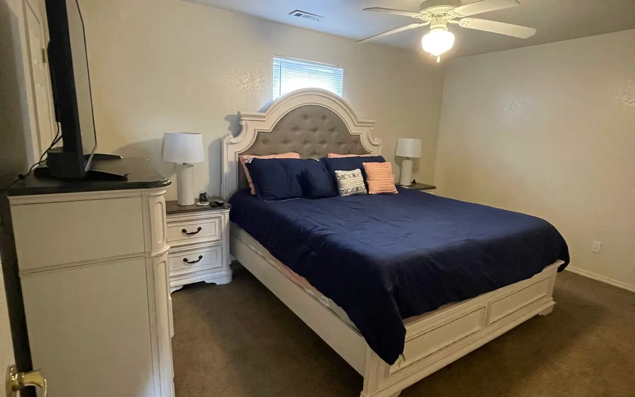 A cozy bedroom featuring a large bed with navy blue bedding, white furniture including a nightstand and dresser, and soft carpet flooring. A television is on a stand to the left, and there are two lamps on the nightstands.