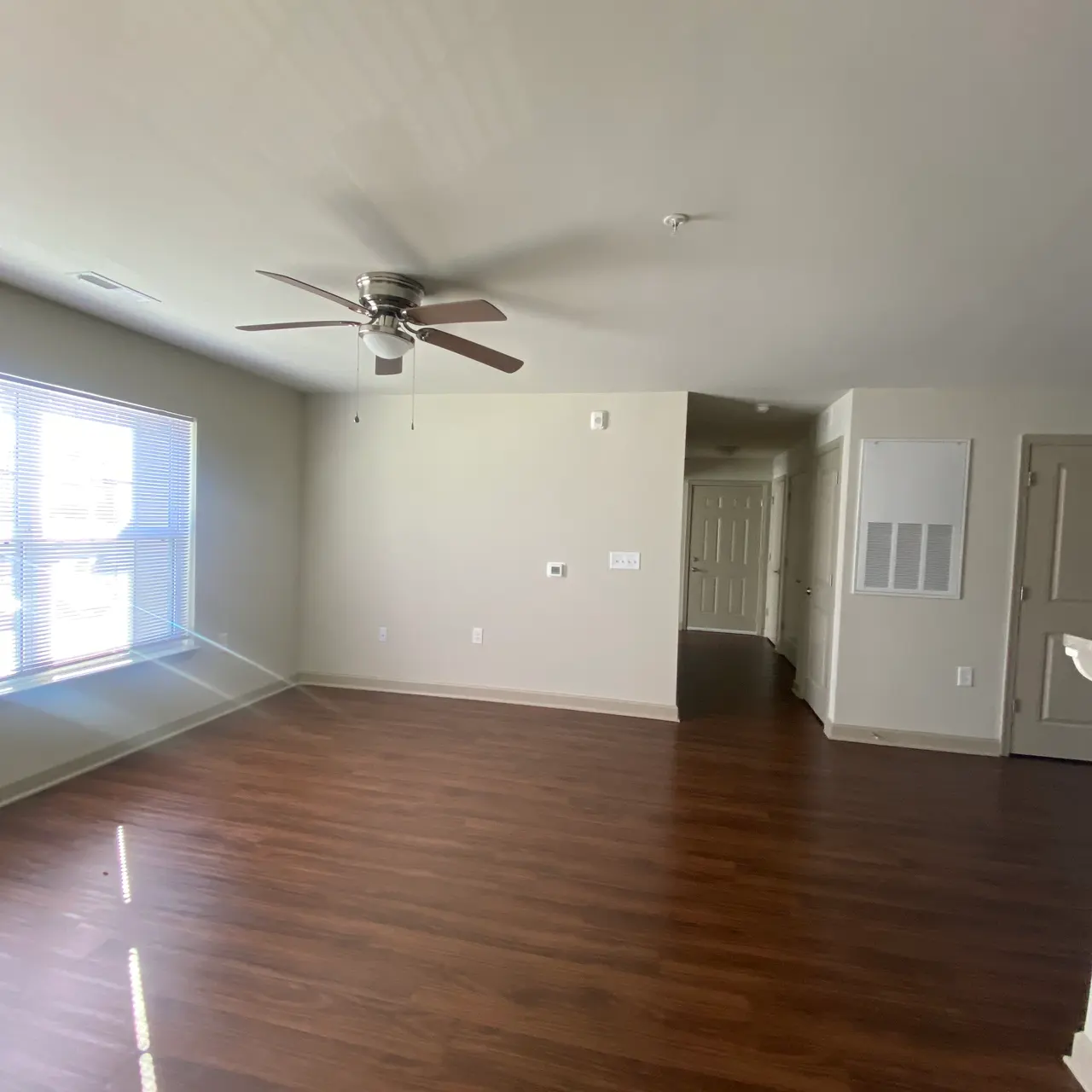 An empty living room showcasing wooden flooring, a ceiling fan, and a large window letting in natural light.