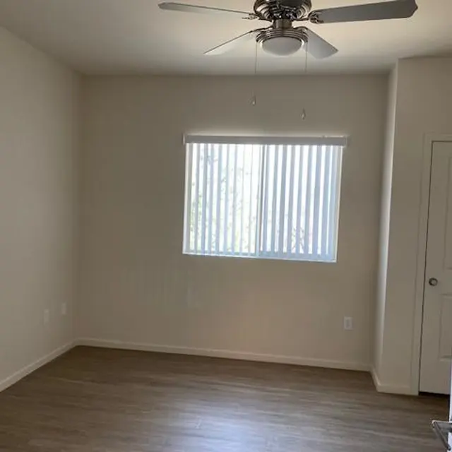 An empty room with light-colored walls, wooden flooring, a ceiling fan, and a window with vertical blinds.