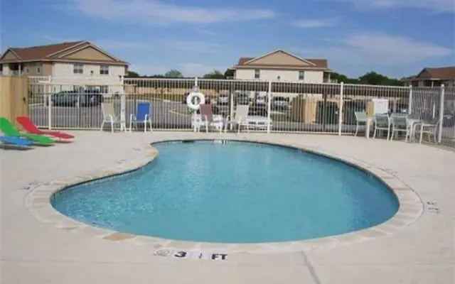 A small, kidney-shaped swimming pool with clear blue water, surrounded by a fence and poolside chairs in bright colors. There are neighboring buildings in the background.